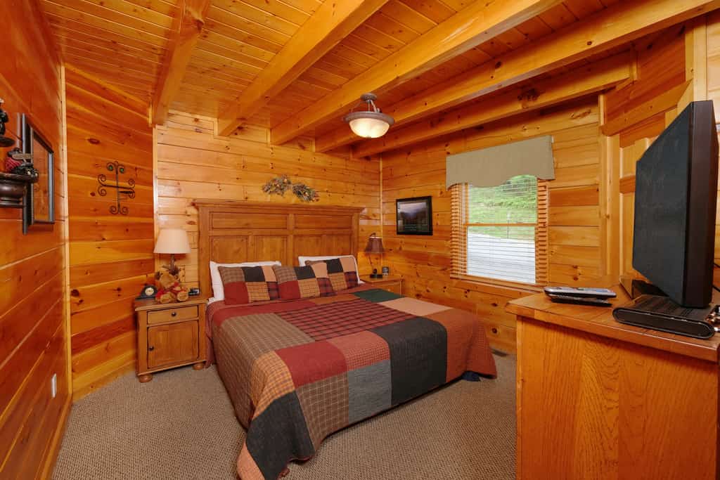 A cozy log cabin-style guest room with a large bed, flat screen TV, and window with a view.