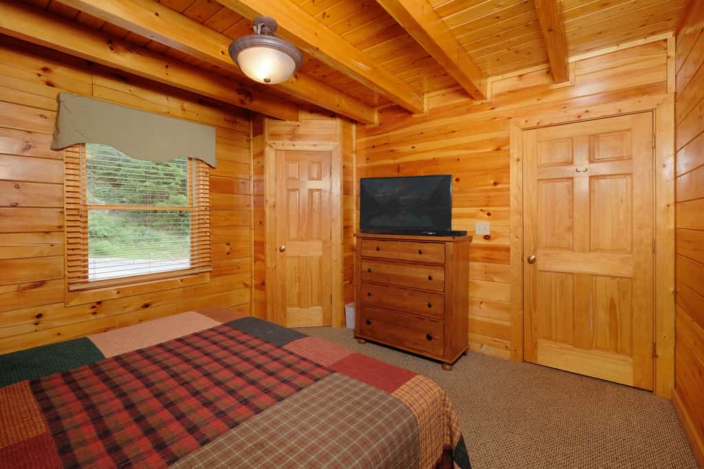 A wide-angle view of a cozy room with wood paneling, a patchwork bedspread, a flat-screen TV on a wood dresser, and a window showing greenery outside.