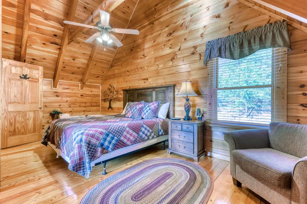 A wide view of a cozy room with wood paneling, a bed, nightstand, armchair, and window.