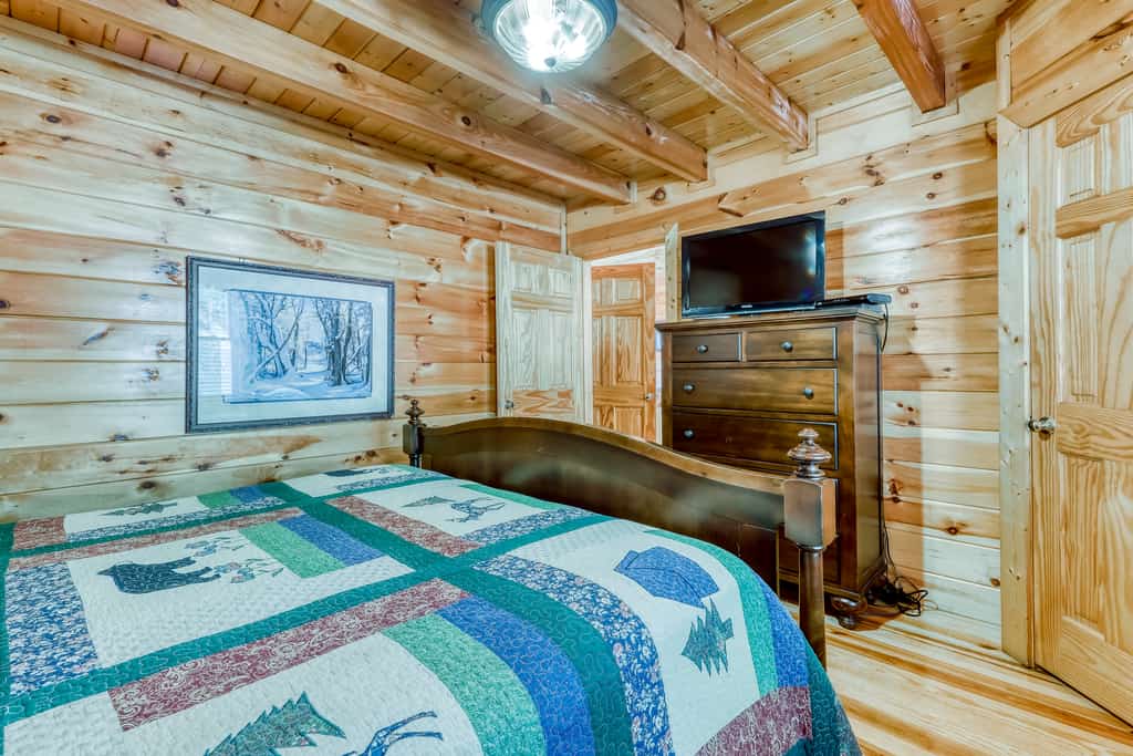 A cozy room with wood paneling, a bed with a patchwork quilt, and a flat-screen TV on a dresser.