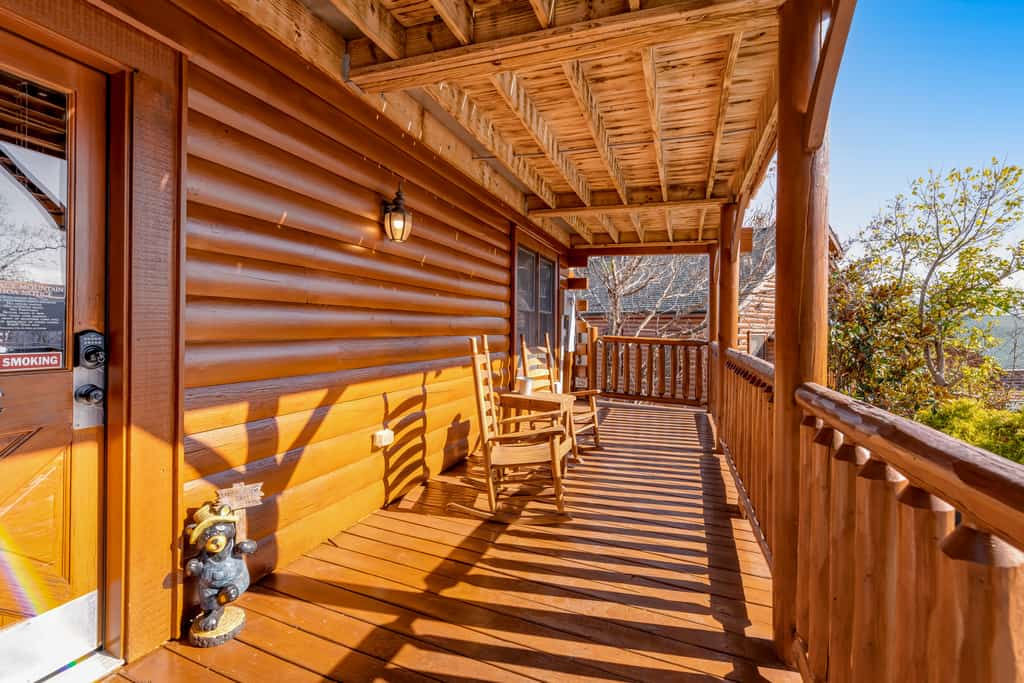 A wooden porch with a door, two chairs, and a railing overlooking trees and a blue sky.