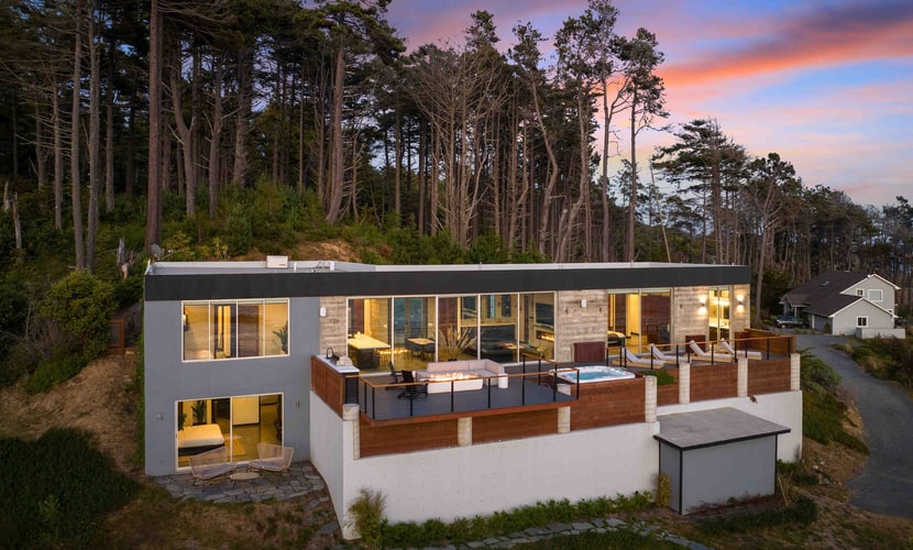 Floor-to-ceiling glass, a private cove, and the Pacific straight ahead. The house is concrete, steel, and wood on 5.25 acres of bluff at Anchor Bay, with 1,800 feet of oceanfront below it and rock islands offshore where the seals bark and, in winter, whales pass close enough to watch from the hot tub. Three suites sleep 8 — two kings and a bunk room. Evenings go to the deck: the 6-person hot tub, the fire pit, the bench at the edge of the bluff, and whatever the sunset decides to do.
