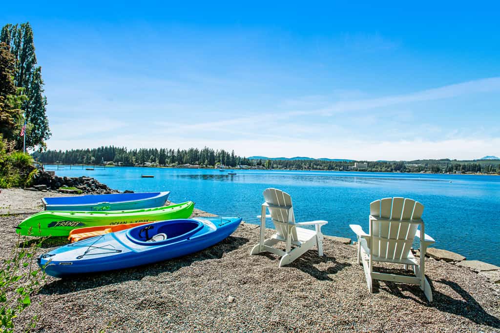 colorful kayaks and other boats line a rocky beach next to two white adirondack chairs