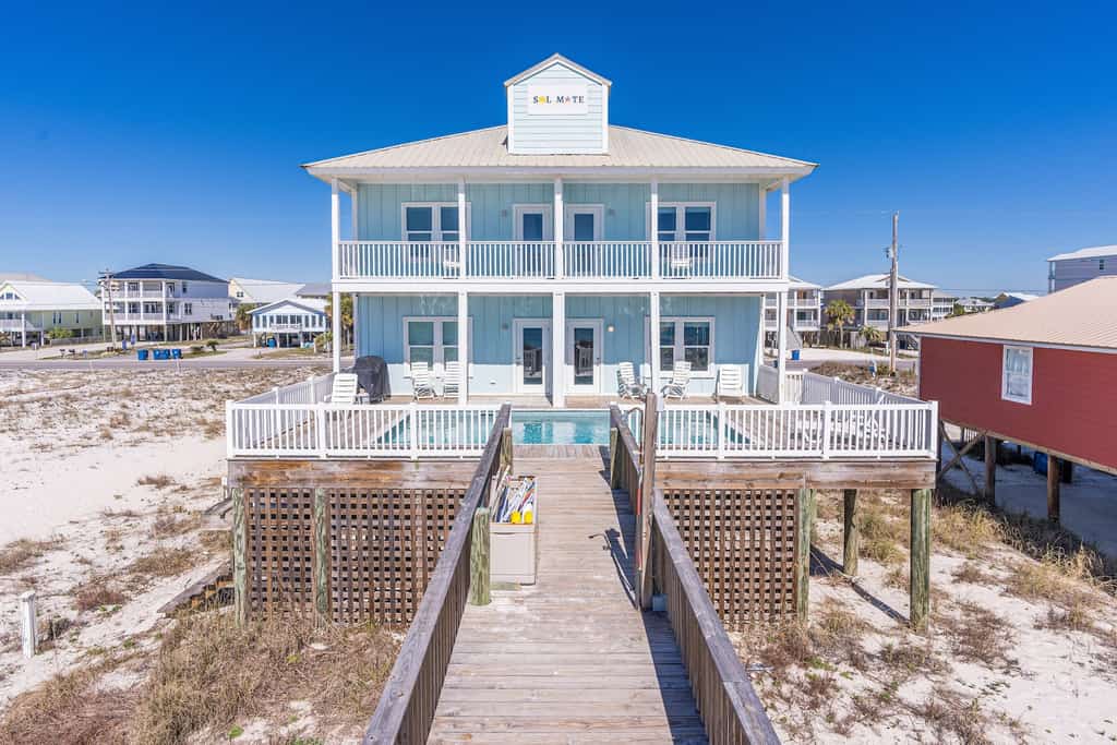 The back pool area with a dock leading up to a blue two story rental in the Gulf Shores.