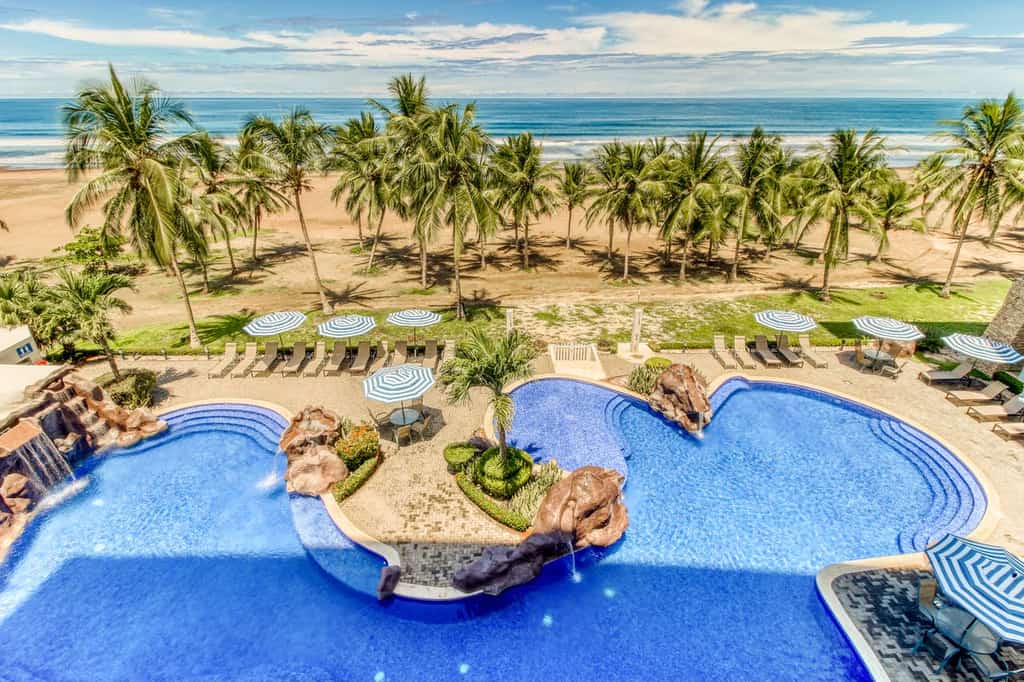 Outdoor pool at a resort with a waterfall near a sandy beach with palm trees.