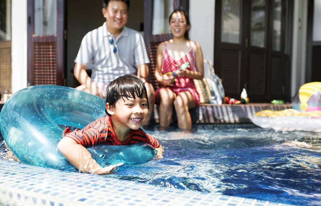 a young boy in an inflatable inner tube swims in a pool with his parents nearby