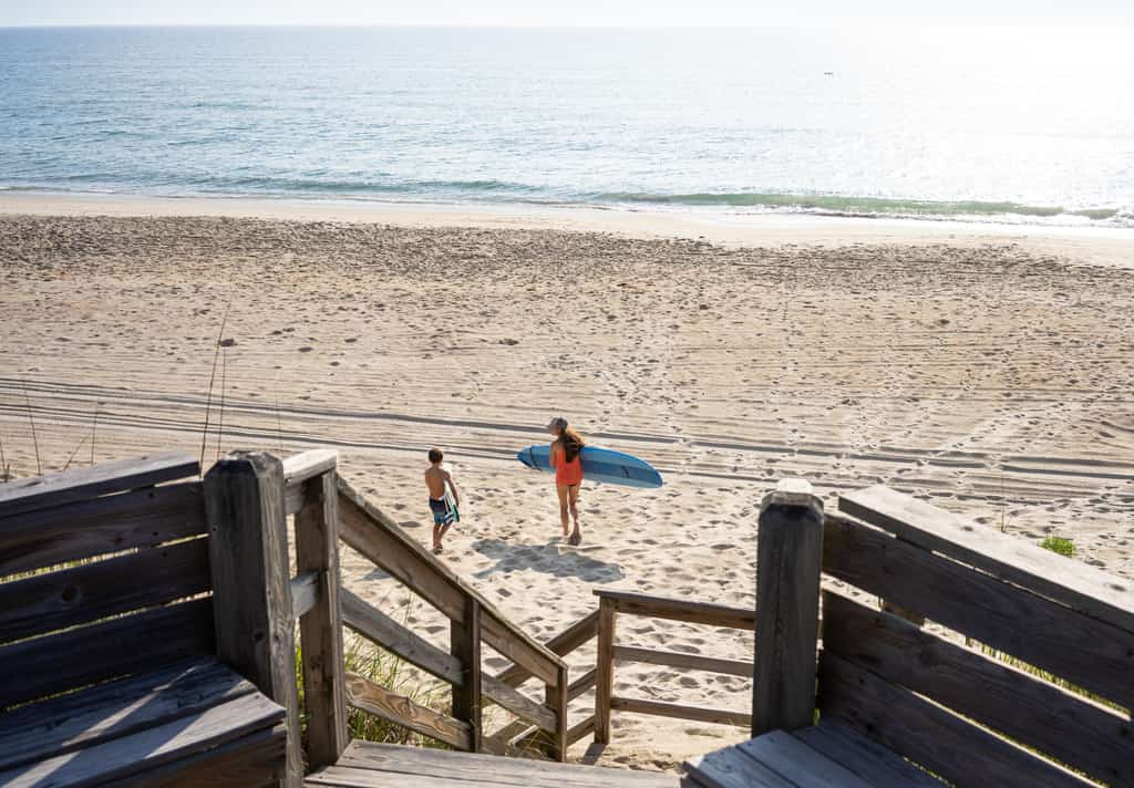A woman and boy carrying surfboards on a sandy beach