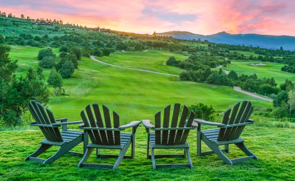 Four adirondack chairs on a well groomed field of grass facing towards a hill with a trail in Beaver Creek, CO
