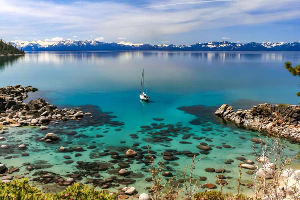 Lone sailboat in crystal clear water with a view of snow capped mountain range at Lake Tahoe
