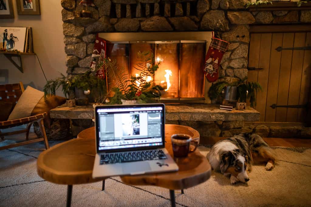 a laptop sits on a coffee table in front of a lit fireplace and a napping dog