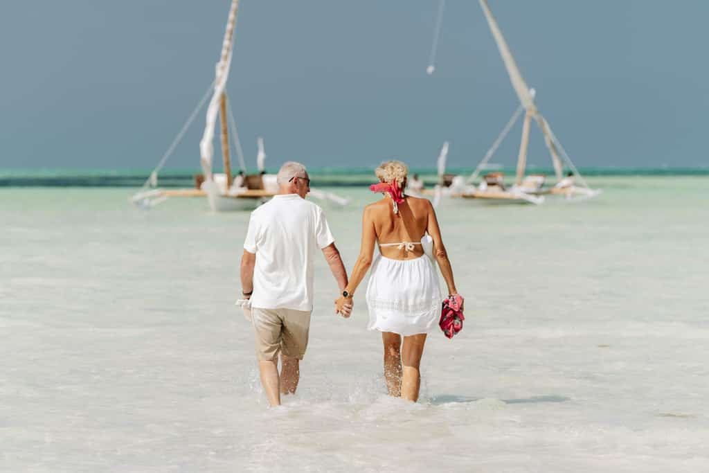A couple holding hands in a beach walking towards two boats.