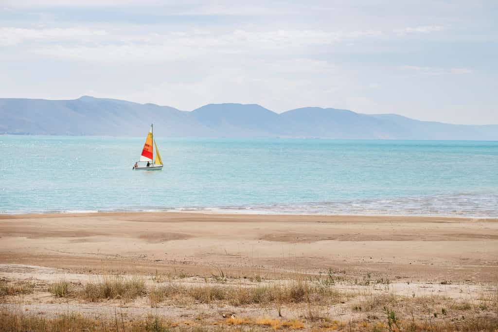view from a sandy beach of a sailboat with two people on board on bear lake in summer