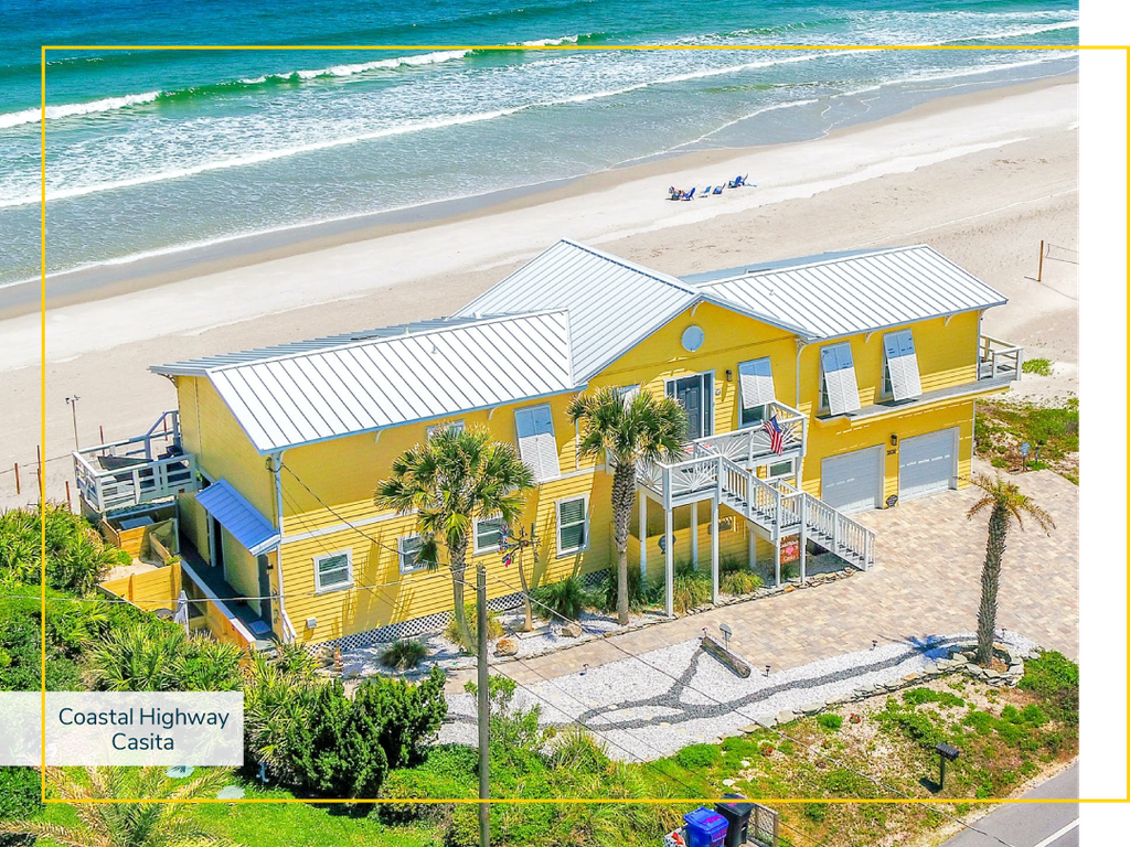 Aerial view of a yellow vacation home on a sandy beach near the shore during the day.