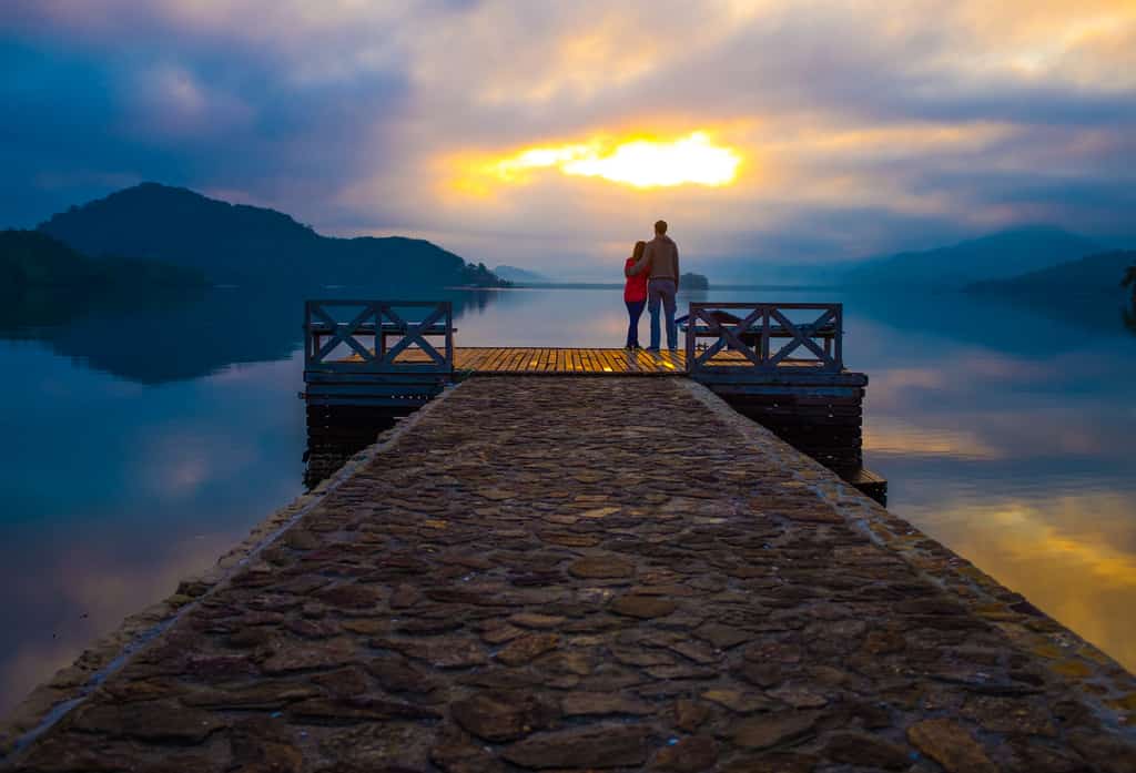 Couple standing on a dock while watching the sunset over a lake.
