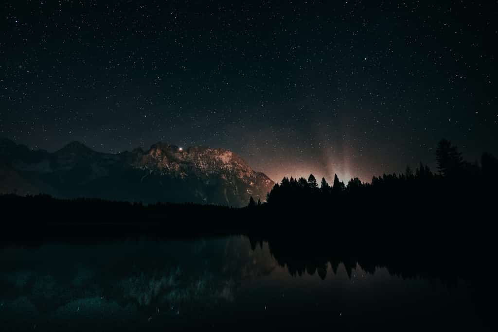 Image of a lake shadowed by a mountain with a bright light and a vast view of memorable star features.
