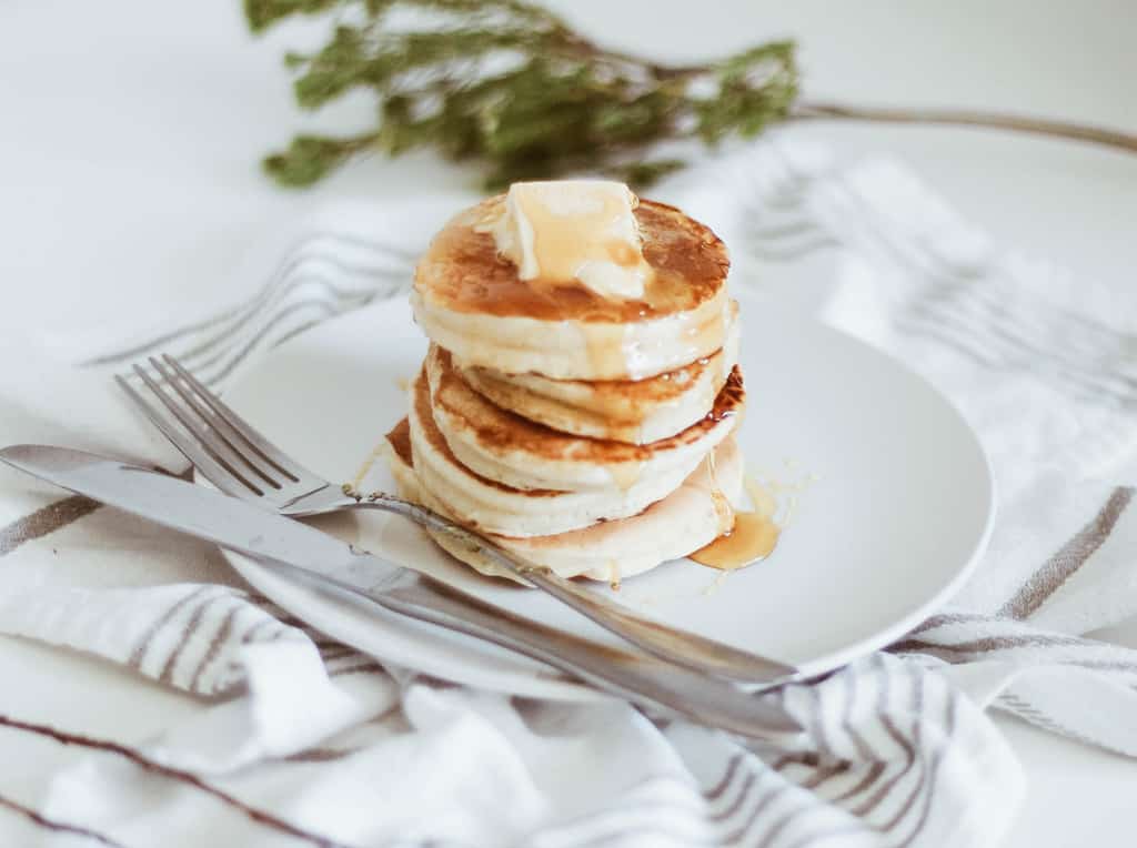 Stacked pancakes with butter and syrup on a white plate with a fork and knife.